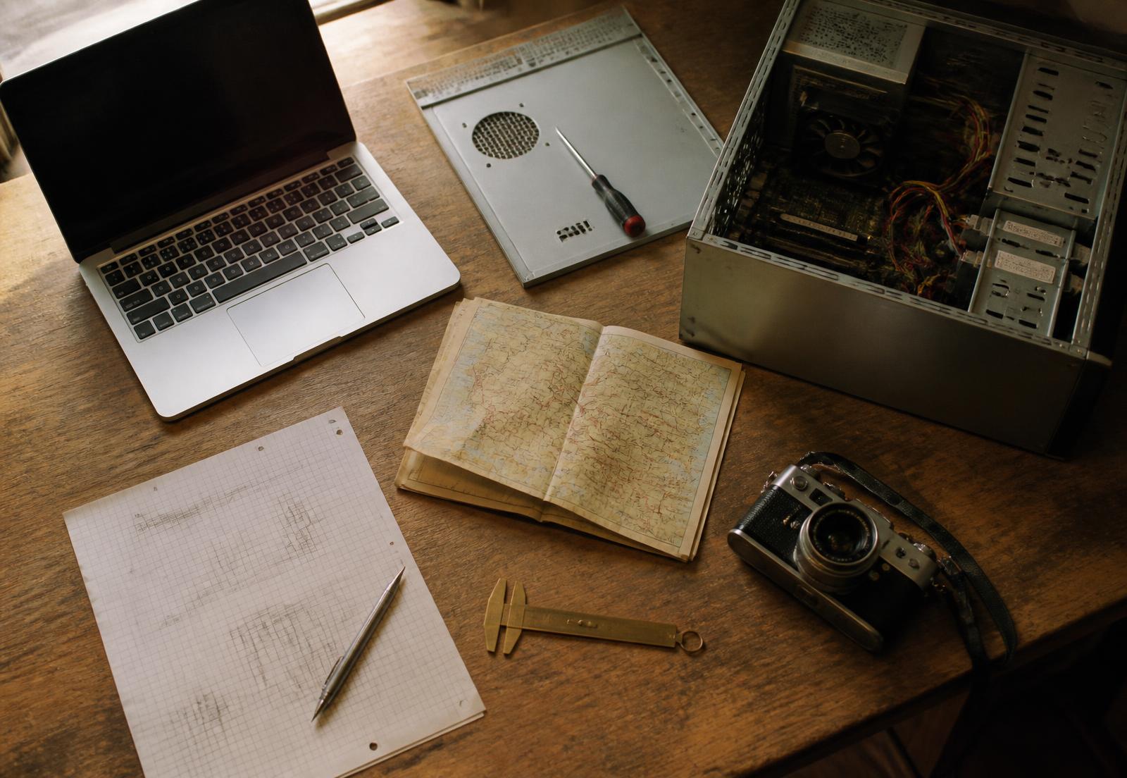 Overhead view of the studio worktable: laptop, opened computer case, screwdriver, folded map, graph-paper sketches, brass caliper and film camera